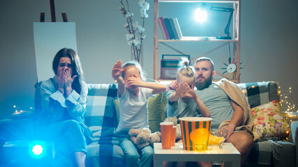 Shocked. Happy family watching projector, TV, movies with popcorn and drinks in the evening at...