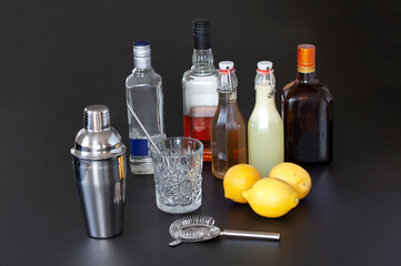 Bar accessories, drink tools and cocktail ingredients including shaker, strainer, glass, different liqueur with homemade syrup bottles and three lemons. Image on black background.