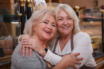 Cheerful senior mother and her mature daughter hugging, enjoying working together at their bakery...