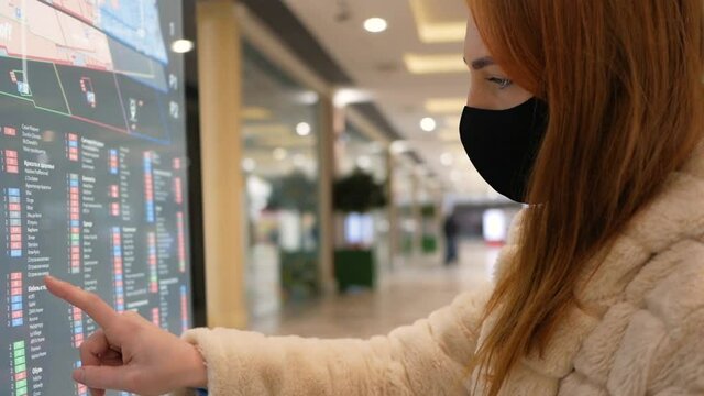 A young red-haired girl in a black mask from Covid-19 examines a map of a shopping center through a tablet