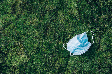 Used blue hygienic medical protective mask on green grass background. Top view. Copy space. Environmental pollution and waste during coronavirus Covid-19 pandemic. Plastic pollution problem