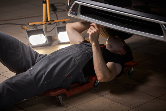 caucasian auto mechanic lying and working under car, necessary instruments on the floor next to him