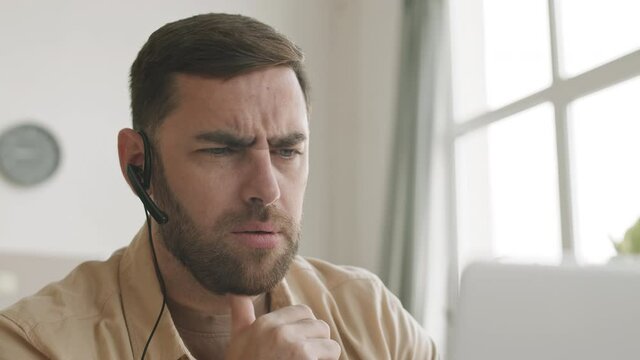 Close-up of male blue-eyed bearded Caucasian person wearing headset in one-ear looking at computer screen talking, frowning, shaking head no, disapproving. Man working from home 