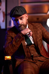 handsome young bearded man in a tuxedo sitting in dark room in fashionable clothing for the festive evening