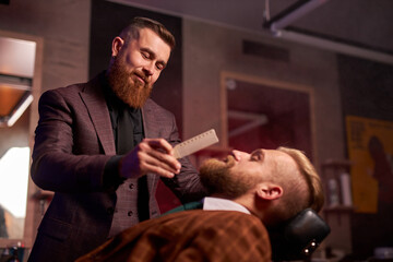 hairstyling process. barber cutting beard of caucasian male with the use of scissors, client sits in an armchair