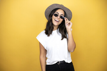 Beautiful woman wearing sunglasses, casual white t-shirt and a hat standing over yellow background smiling