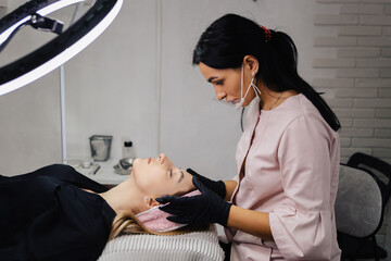 The girl lies with closed eyes on the couch. A master cosmetologist in a medical gown, mask and black gloves checks the proportions and sketch of the eyebrow on the girl's skin before microblading.