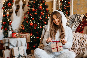 Obraz na płótnie Canvas Young smiling woman picks a box with a Christmas present in her hands, relaxing at home, enjoying Christmas. A beautiful girl at home, sitting near the Christmas tree. Gift for the new year