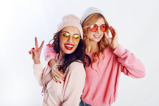 Indoor studio image of two girls, happy friends in stylish pink clothes and hat spelling funny  the together. White background. Trendy hat and glasses.Showing peace.