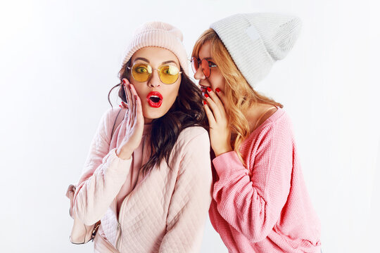 Two cheerful young friends in perfect mood having time together on white isolate background. Positive girls wearing pink  cozy sweaters and pullover, trendy hat , cute glasses.