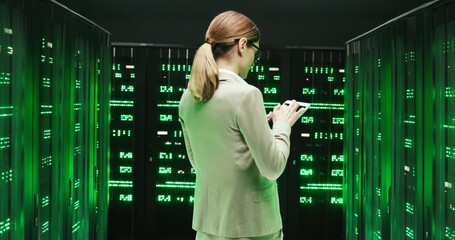 Rear of Caucasian young woman with walking among servers and checking work of big data processors. Back view Female analytic in data storage strolling in information security office with tablet device