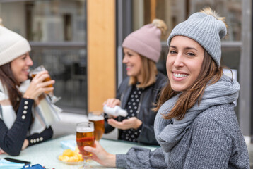 Young smiling women wearing winter clothes drinking beer on a bar. One woman is disinfecting her hands with gel. One woman is looking at camera. New normal concept