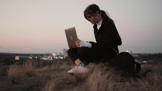 Business woman typing on a laptop keyboard in a sunset time outdoors on hills in industrial zone near wind energy farm. Spirit of youth freedom in independent internet business.