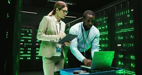 African American man with laptop computer and Caucasian woman with tablet device talking and working on software of big data center. Mixed-races male and female analytics colleagues. Digital security.