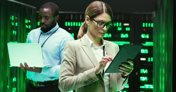 Caucasian woman tapping on tablet device and checking big data security. Female analytic in data storage working on information hardware. African American male co-worker with laptop on background.