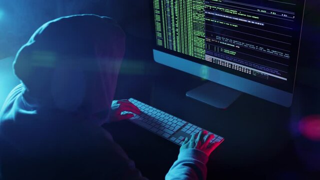 Back view of hacker programmer wearing a hood, typing very fast intelligible code in a computer screen. Mysterious woman hacking in a dark smoky room. Concept dark internet, cyber security.