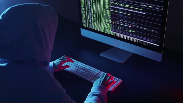 Back view of hacker programmer wearing a hood, typing very fast intelligible code in a computer screen. Unrecognizable woman hacking in a dark room. Concept dark internet, cyber security.
