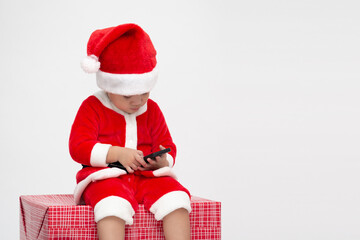 Asian baby boy with Santa Claus suit holding mobile phone and sitting on gift box isolated on white background