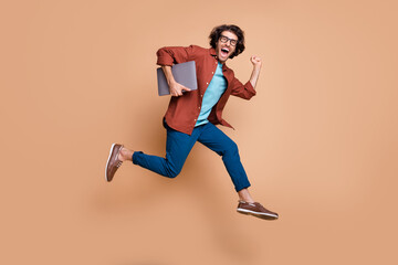 Photo portrait full body view of screaming guy in casual brown shirt running with laptop in arm jumping up isolated on pastel beige colored background