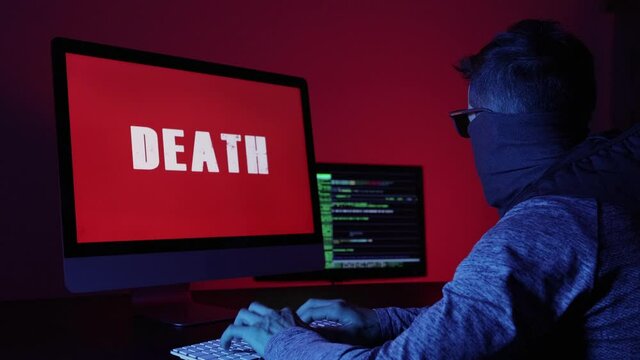 Man with a covered face in front of two computer screens. Criminal hacker programming intelligible codes and death. Computer crime concept.
