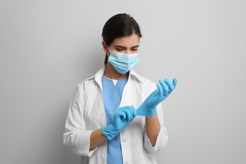 Doctor in protective mask putting on medical gloves against light grey background