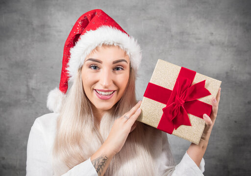 Surprised beautiful blond woman with wears christmas santa claus hat - cap and holding red gift box - present with ribbon, isolated on gray background