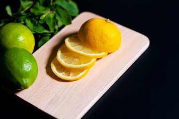 On a wooden Board is a sliced lemon. Next to it are whole fruits with mint leaves on a dark background. The view from the top.