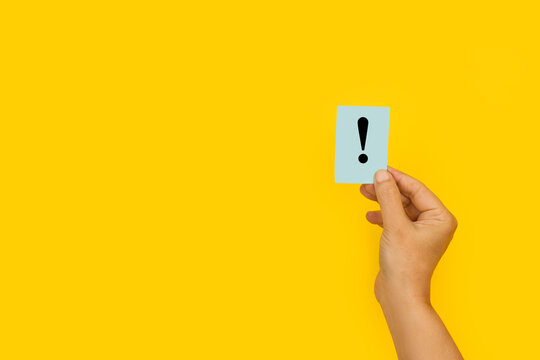 Mano de mujer sosteniendo un papel con el signo de exclamación sobre el fondo amarillo brillante liso y aislado. Vista de frente. Copy space 