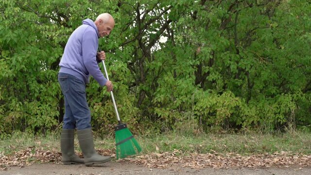 old grey haired gardener man sweeping dry foliage on asphalt road side view zoom out