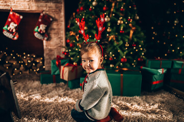 Little girl sitting in front of a decorated Christmas tree. Against the background of a Christmas tree with boxes in green packaging with a bow and a fireplace. Christmas mood