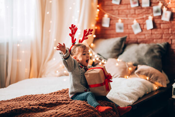 A little girl holding a large gift in a paper box on a large bed. The child has antlers on his head. Christmas mood