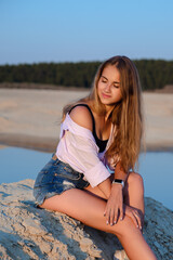 A young and beautiful girl sits on the sand with her eyes closed and enjoys the setting sun.