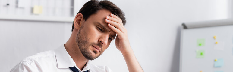Fototapeta premium Exhausted businessman with migraine holding hand on forehead, while looking away on blurred background, banner