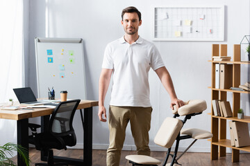 Front view of positive physiotherapist looking at camera while leaning on massage chair in office