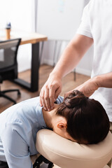Obraz na płótnie Canvas Massage therapist massaging neck of brunette businesswoman sitting on massage chair on blurred background