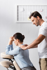 Smiling masseur doing seated massage of businesswoman shoulders in office on blurred background
