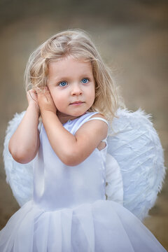 Beautiful chubby blonde angel with blue eyes in a white ball gown with white feather wings. Vertical photo.