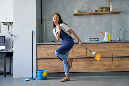 Happy worker. Full length shot of cheerful young woman, cleaning lady pretending to sing a song, holding broom while cleaning the floor, doing household chores