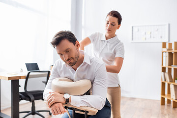 Female massage therapist massaging back of businessman sitting on massage chair in office on blurred background