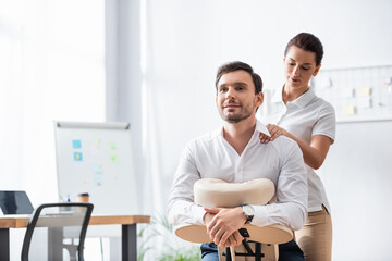 Masseuse massaging shoulder of smiling businessman sitting on massage chair in office on blurred background - Powered by Adobe