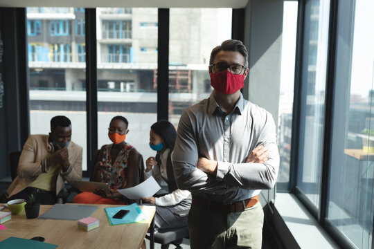 Portrait of caucasian man wearing face mask standing with arms crossed at modern office