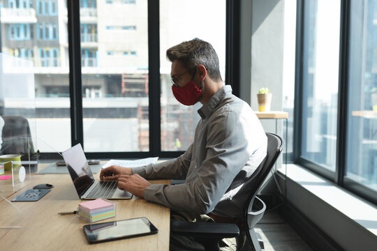 Caucasian man wearing face mask using laptop while sitting on his desk at modern office