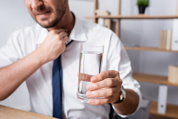 Cropped view of businessman with hand on neck, holding glass of water, while sitting at workplace on blurred background