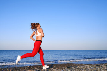 Positive sportive woman on a summer morning jogging on the beach in red leggings on the background of the sea coast