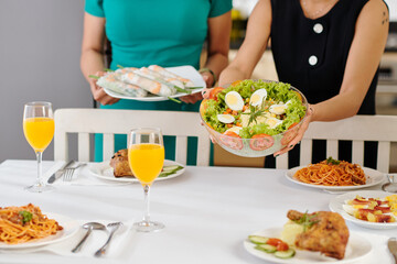 Women putting bowl of delicious vegetable salad and spring rolls on dinner table
