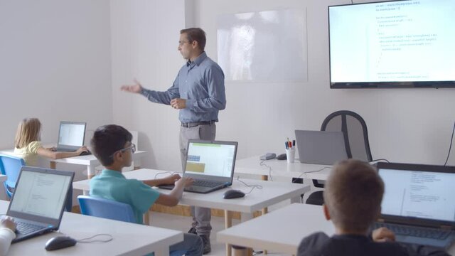 Male computer science teacher explaining lesson to children in class. Wide shot. Digital education concept