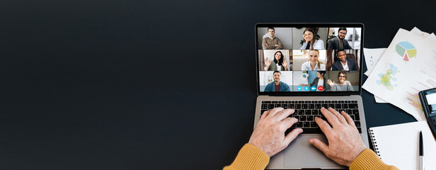 Top view of laptop screen with video conference participants and mature man's hands on keyboard, laptop stands on a black desktop. Copy space