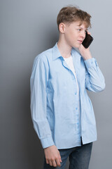 Young guy 14-16 years old in a blue shirt talking on the phone on a gray background