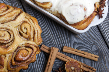 Baked cinnabons in shape. Some are covered with glaze. Stand on black composite boards. Nearby are cinnamon sticks. Close-up shot.