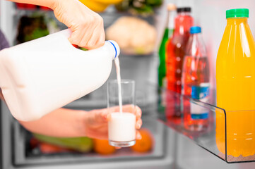 A girl pours milk from a bottle into a glass against the background of an open refrigerator with food. Close-up, small depth of field bright lighting